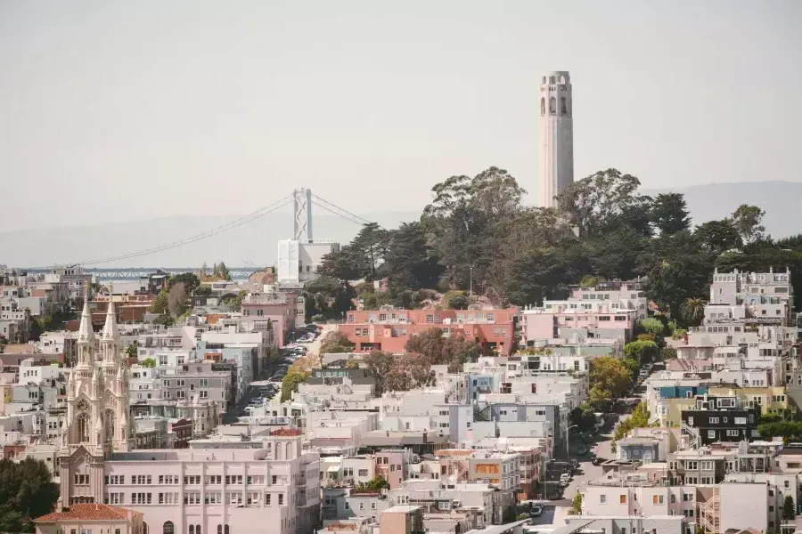 샌프란시스코의 코이트 타워(Coit Tower)는 배경에 베이 브리지(Bay Bridge)가 있고 전경에 집들로 뒤덮인 언덕이 있는 사진입니다.